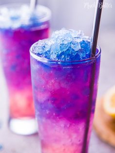 two glasses filled with purple and blue liquid next to an orange slice on a table