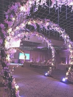 an archway decorated with purple flowers and greenery is lit up at night for a wedding reception