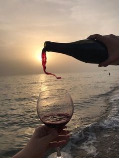 someone is pouring wine into a glass on the beach at sunset or sunrise, while another person holds out their hand