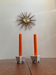 two orange candles sitting next to each other on top of a wooden table in front of a clock