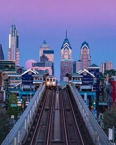 a train traveling over a bridge with tall buildings in the background at dusk, as seen from across the river