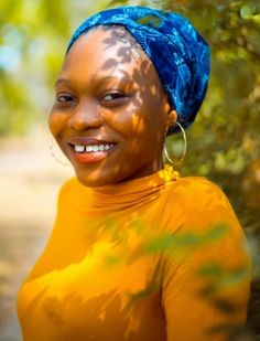 a woman with blue hair wearing a yellow shirt and smiling at the camera while standing in front of some trees