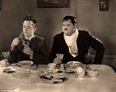 two men sitting at a table with food in front of them, one holding a knife and the other eating