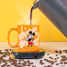 a mickey mouse coffee mug is being poured into a cup with some coffee beans scattered around it