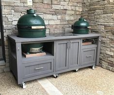 two big green egg grills sitting on top of a gray cabinet next to a stone wall