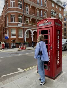 a woman standing next to a red phone booth on the side of a road in front of a tall brick building