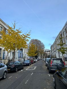 cars parked on the side of a street next to tall white buildings and trees with yellow leaves