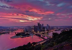 the city is lit up at night with pink and purple clouds in the sky above it