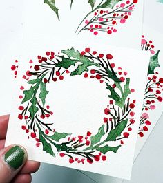 two christmas cards with holly wreaths painted on them, one being held up by a person's hand