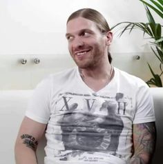 a man with long hair sitting on a couch in front of a potted plant