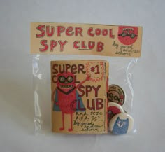 two badges in plastic packaging on a white surface with the words super cool spy club