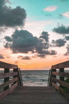 a wooden bridge leading to the ocean at sunset with clouds in the sky above it