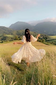 a woman in a white dress is walking through tall grass with mountains in the background