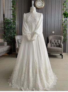 a white wedding gown with long sleeves and lace on the skirt is displayed in front of a clock