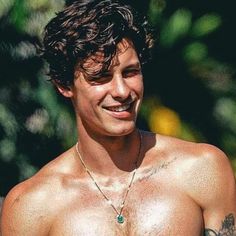 a shirtless man with tattoos on his chest smiles at the camera while standing in front of some trees