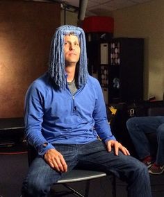 a man with long blue hair sitting on a chair in front of another man who is wearing a wig