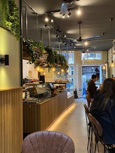 people sitting at tables in a restaurant with plants hanging from the ceiling and lights above them