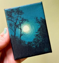 a hand holding up a small painting with trees and the moon in the sky behind it