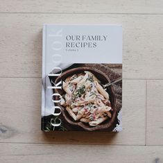 the cookbook is sitting on top of a wooden table next to a bowl of pasta