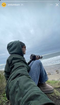 a man sitting on top of a grass covered field next to the ocean holding a camera