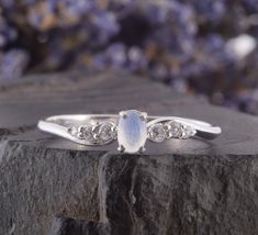 an oval shaped white stone ring with three small diamonds on top of it, sitting on a rock in front of purple flowers