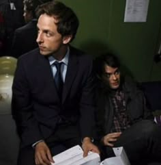 two men sitting next to each other in front of a green wall and one man holding papers