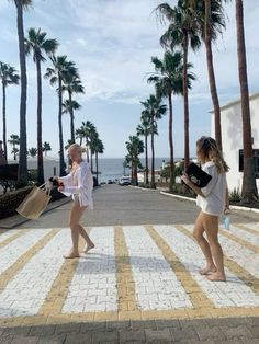 two young women walking down a sidewalk next to palm trees and the ocean in the background