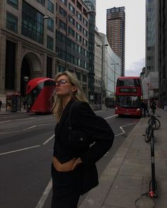 a woman standing on the side of a road in front of tall buildings with red double decker buses