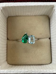 an engagement ring with two pear shaped green stones in a white box on carpeted surface