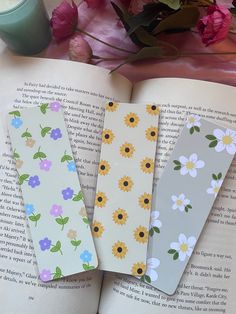 three bookmarks sitting on top of an open book next to flowers and a candle