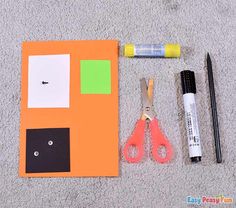 scissors, markers, and glue are laid out on the floor next to an orange piece of paper