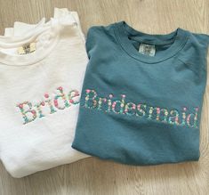 two t - shirts with embroidered words on them sitting on a wooden floor next to each other