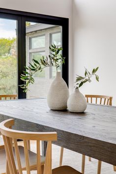 two white vases sitting on top of a wooden table next to a window with an olive branch in it