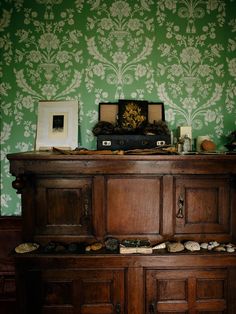 an old wooden cabinet in front of a green wallpapered room with pictures on it