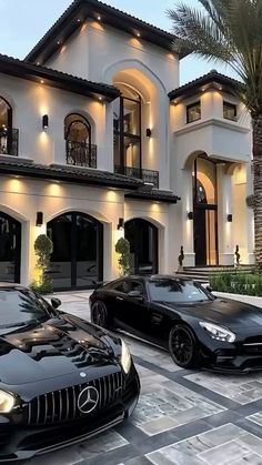 two black sports cars are parked in front of a large white house with palm trees