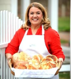 a woman in an apron holding a basket full of baked goods on the side of a white picket fence