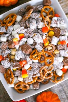 a white square bowl filled with candy corn and pretzel treats next to mini pumpkins