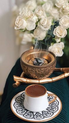 a cup and saucer on a table with white roses in the background, next to a gold - plated teacup