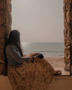 a woman sitting on a window sill looking out at the ocean and beach in front of her