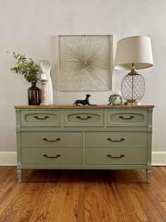 a green dresser sitting on top of a hard wood floor next to a lamp and painting