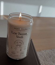 a lit candle sitting on top of a wooden table next to a glass container with the label june twenty sixth