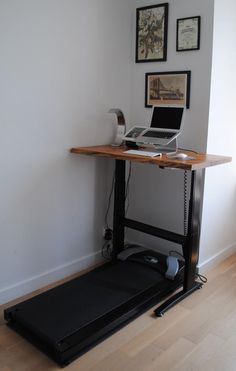 a laptop computer sitting on top of a wooden desk next to a treadmill in front of a white wall