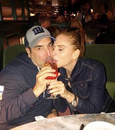 a man and woman sitting at a table kissing while drinking from wine glasses in front of them