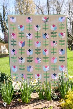 a quilted wall hanging on the side of a tree in a park with flowers