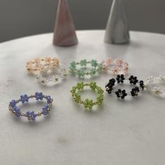 six different colored beaded rings sitting on top of a white table next to a vase