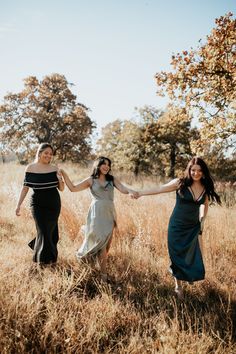 three women in long dresses holding hands and walking through tall grass