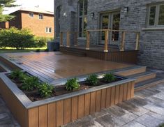 a wooden deck surrounded by plants in front of a brick building with trees and bushes