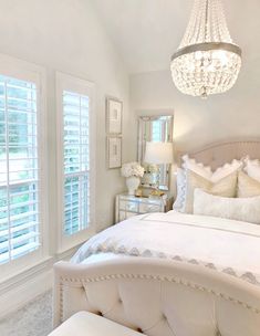 a white bedroom with a chandelier, bed and two windows that have shutters on them