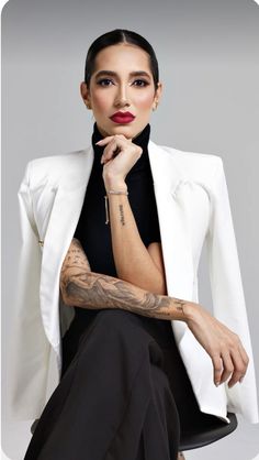 a woman in a white jacket and black dress sitting on a chair with her hand under her chin