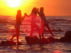 three girls are standing on rocks in the water with their arms around each other as the sun sets behind them